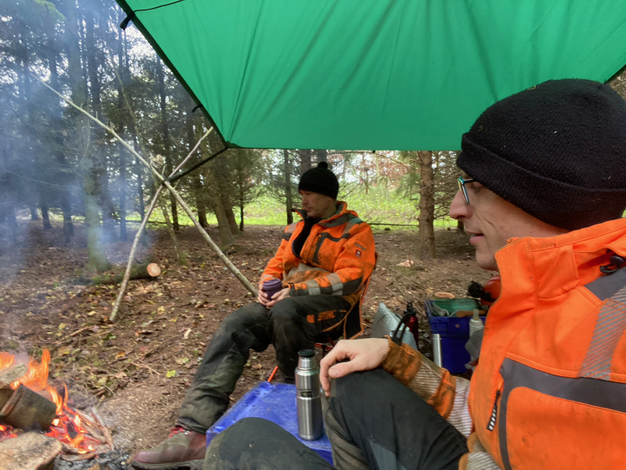 two people sitting next to a fire wearing work clothes in a woodland setting