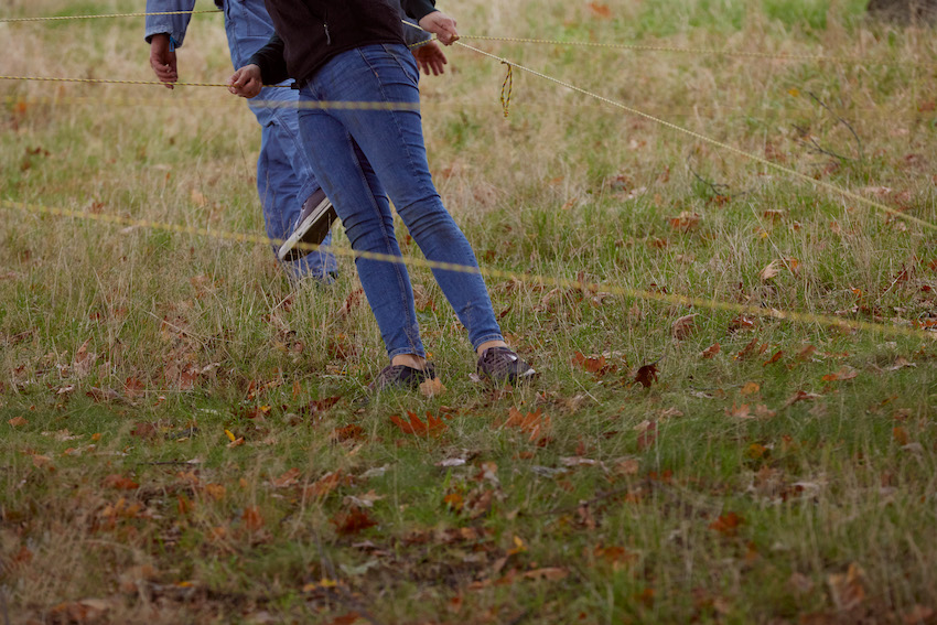 two people standing on grass, leaning to one side while holding horizontal strings