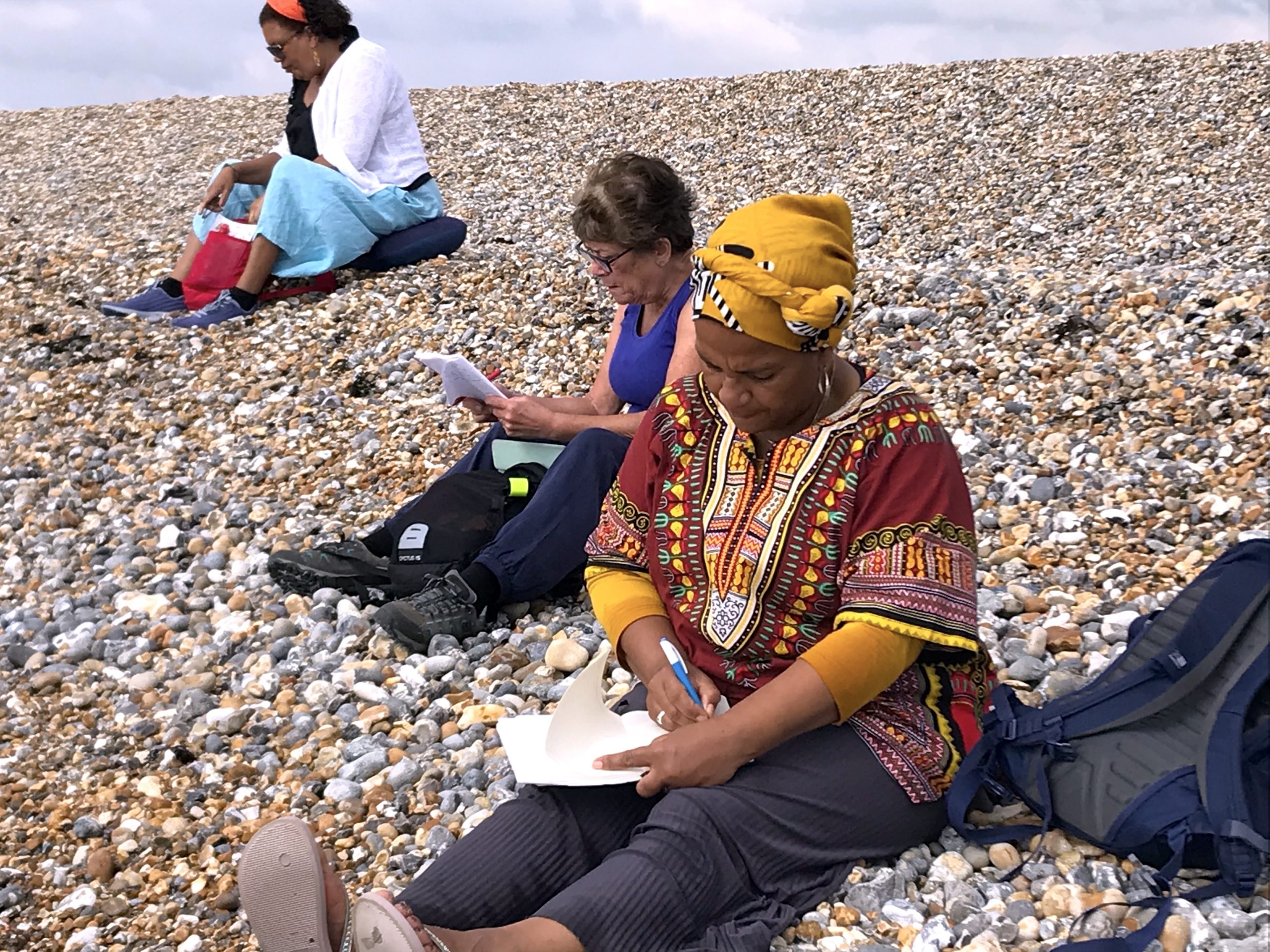 Three women sitting on a pebbly beach, writing