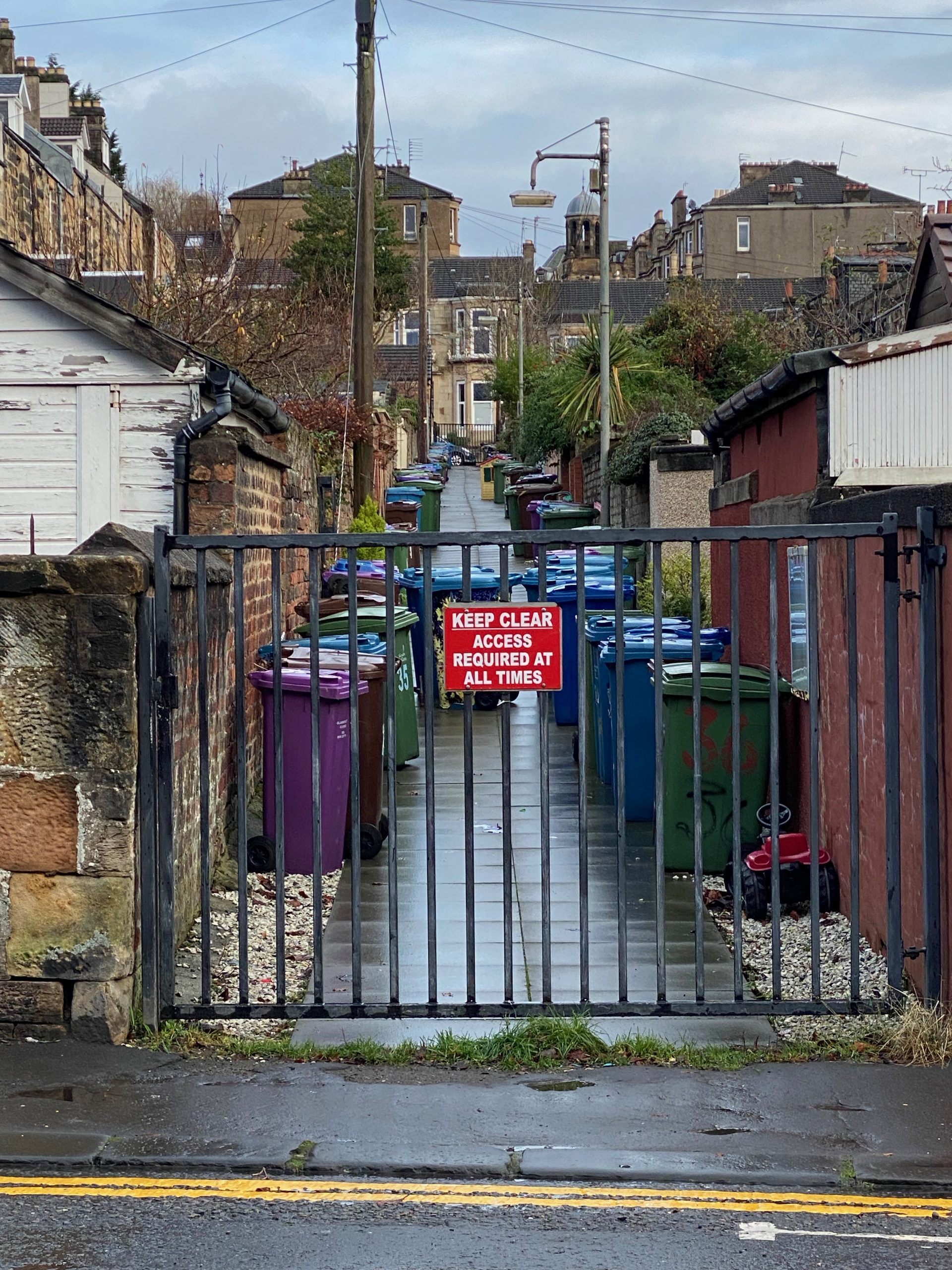 Gated lane between buildings filled with bins. There is a 'keep clear' sign on the closed gate.